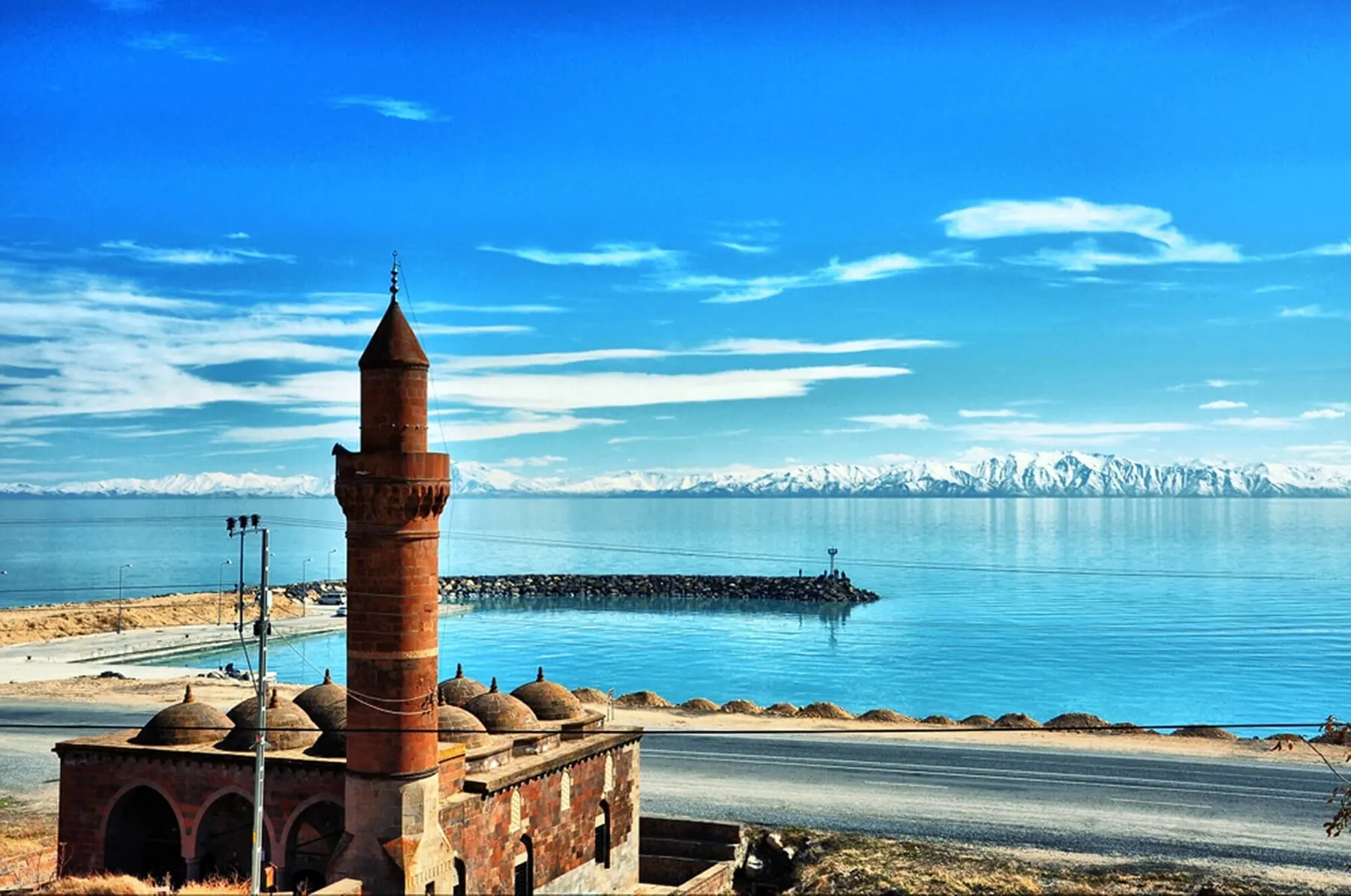 A stone mosque and minaret on the Adilcevaz shore, with blue Lake Van and snowy mountains on the opposite shore.