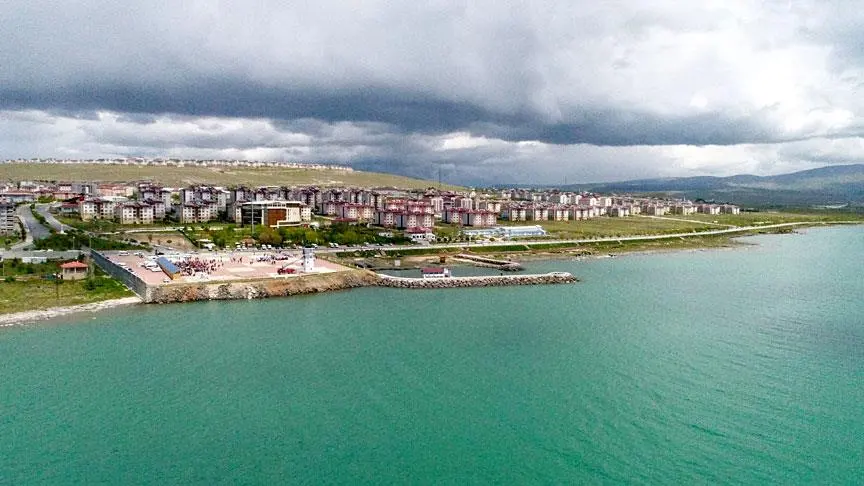 Bulutlu havada gölden sahile doğru bakış; beyaz fener, meydan ve kıyıdaki konutlar